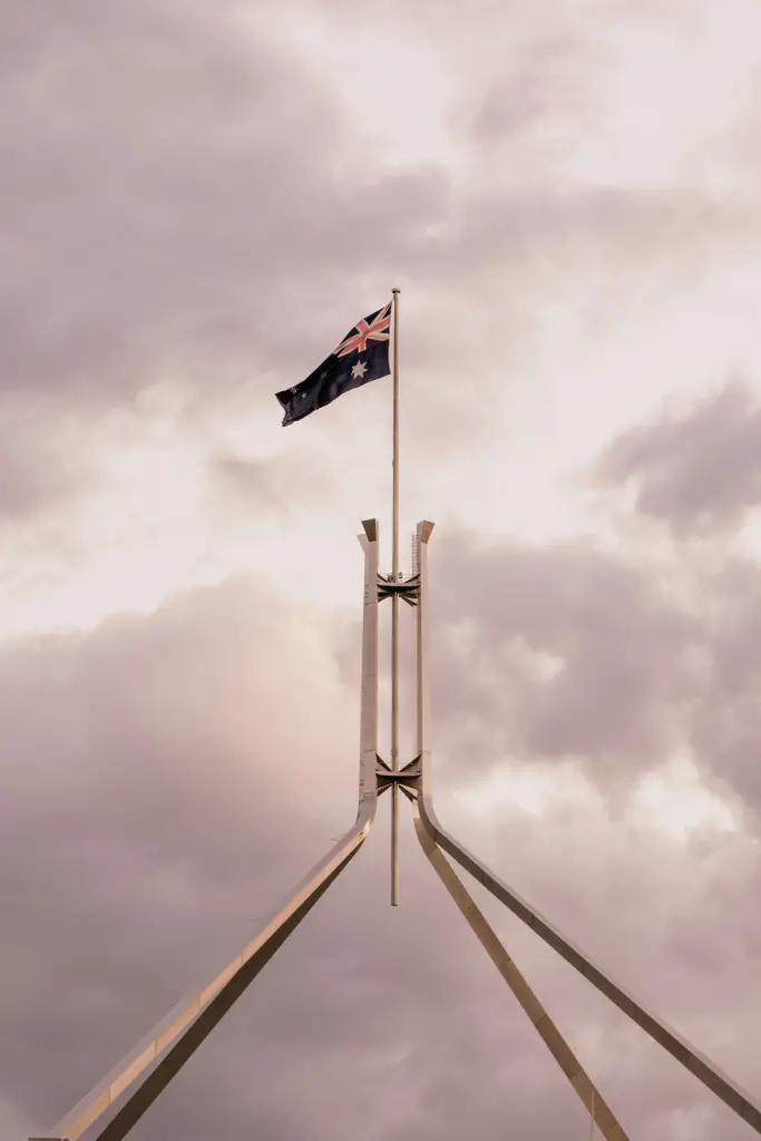 The Australian flag flying high over Parliament House in Canberra with a dramatic cloudy sky background.